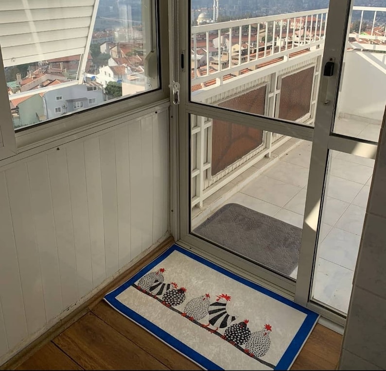May include: A white door with a blue and white patterned doormat featuring a line of black and white chickens. The doormat is on a wooden floor.