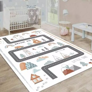 Puede incluir: Una alfombra blanca para niños con un diseño de carretera y animales de dibujos animados. La alfombra presenta una carretera gris con ilustraciones de un coche, conejo, zorro, árboles, montañas y el texto "DREAM BIG" y "LITTLE ONE".