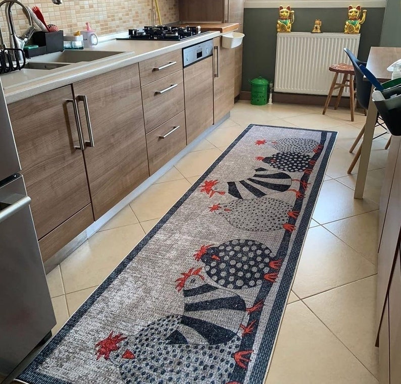 May include: A long, rectangular, gray and white kitchen rug with a black and white chicken design. The rug has a black border with a white stripe.