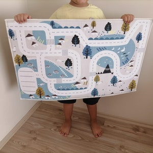 Puede incluir: Una alfombra de juego blanca y azul con un dise&ntilde;o de carretera y ciudad. La alfombra presenta un camino sinuoso, casas, &aacute;rboles y una monta&ntilde;a.
