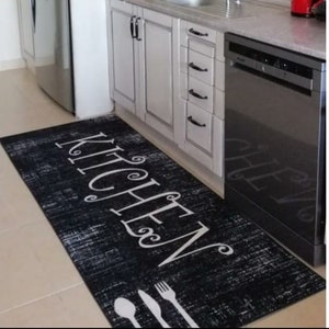 Op de afbeelding: Een rechthoekig zwart keukenkleed met de witte tekst "KITCHEN". Het kleed heeft een distressed design en witte silhouetten van bestek aan de onderkant. Het kleed ligt voor een aanrecht en een vaatwasser.