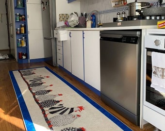 Può includere: Una cucina bianca con elettrodomestici in acciaio inossidabile, tra cui una lavastoviglie, un frigorifero e un forno. La cucina ha armadi bianchi con finiture blu e un piano di lavoro bianco. Un tappeto a motivi bianchi e neri con accenti rossi è sul pavimento.