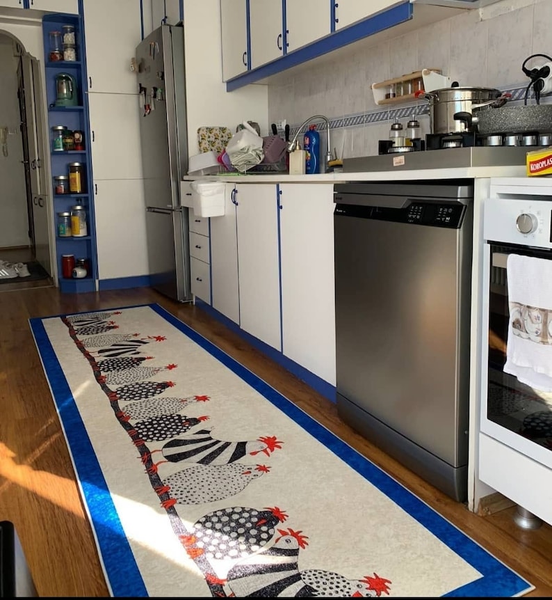 May include: A white kitchen with stainless steel appliances, including a dishwasher, refrigerator, and oven. The kitchen has white cabinets with blue trim and a white countertop. A white and black patterned rug with red accents is on the floor.