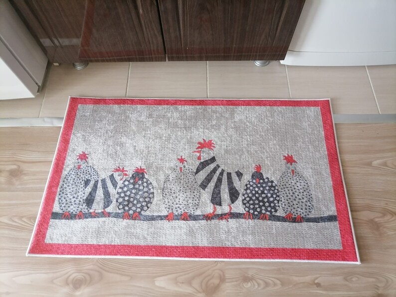 May include: A gray and red rectangular rug with a white border features a design of seven black and white chickens with red combs. The chickens are lined up in a row and appear to be walking on a branch.