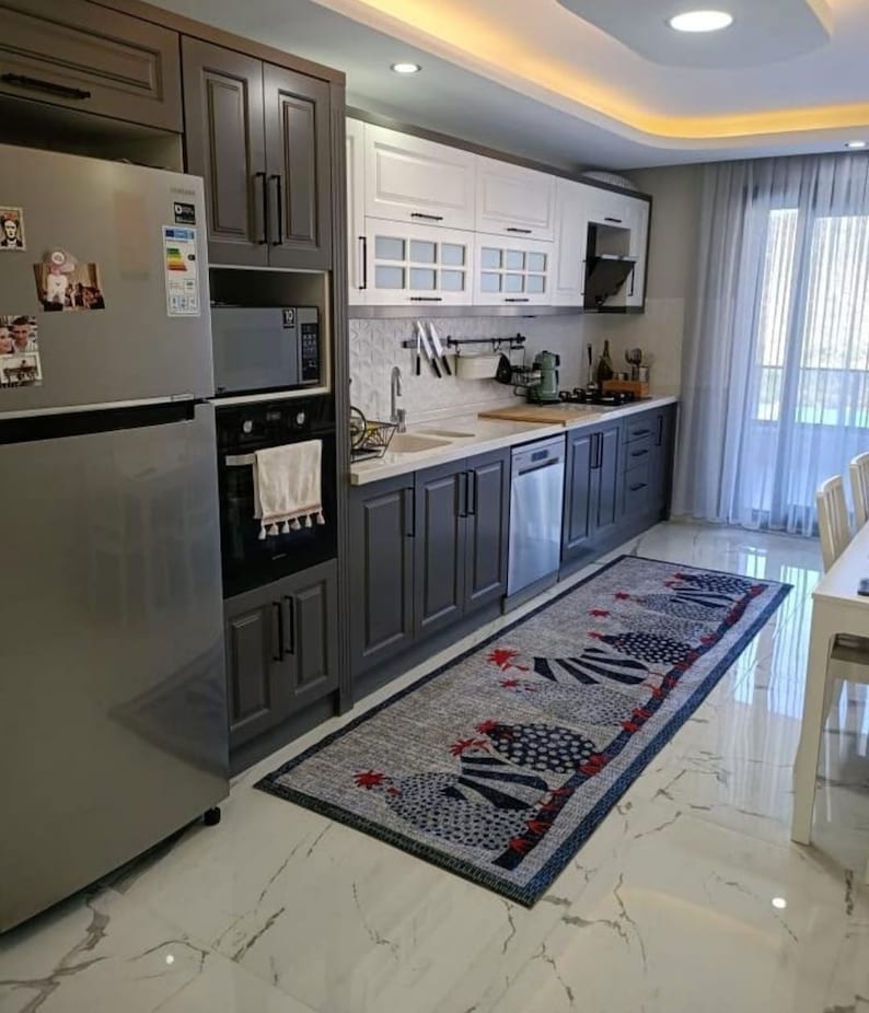 May include: A modern kitchen with dark gray cabinets, white cabinets with glass doors, stainless steel appliances, and a white tiled backsplash. A blue and white rug with a red rooster design is on the floor.