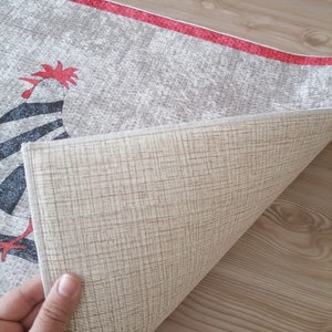 May include: A beige rug with a red border and a black and white rooster design. The rug is folded in half to show the beige backing.