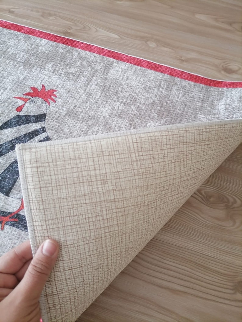 May include: A beige rug with a red border and a black and white rooster design. The rug is folded in half to show the beige backing.
