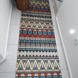 Puede incluir: Una alfombra rectangular estampada con un diseño geométrico en tonos rojos, azules, crema y negro. La alfombra presenta patrones repetidos de triángulos, óvalos y líneas, creando un diseño vibrante. La alfombra está colocada sobre un suelo embaldosado.