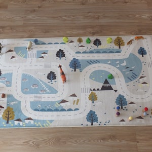 Puede incluir: Una alfombra de juego blanca y azul con una carretera e ilustraciones de animales. La alfombra presenta una variedad de animales de juguete, incluyendo una jirafa, un tigre, una vaca y caballos. Tambi&eacute;n hay clavijas de madera de colores en la alfombra.