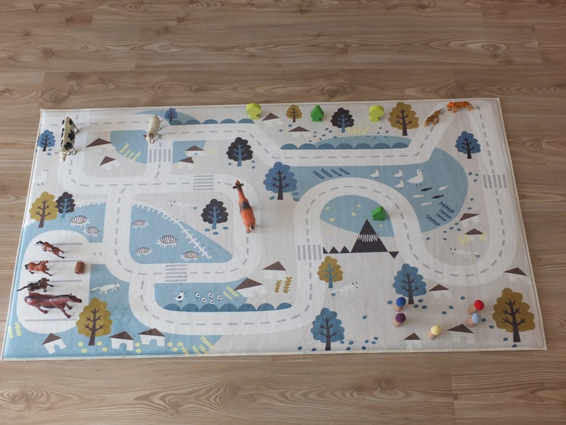 Puede incluir: Una alfombra de juego blanca y azul con una carretera e ilustraciones de animales. La alfombra presenta una variedad de animales de juguete, incluyendo una jirafa, un tigre, una vaca y caballos. Tambi&eacute;n hay clavijas de madera de colores en la alfombra.