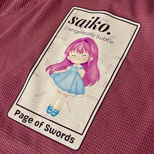 May include: A mauve waffle-knit shirt featuring a graphic of a cartoon girl with pink hair and a blue dress. The graphic is labeled "saiko. Dangerously Subtle." and "Page of Swords."