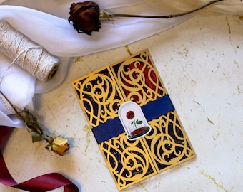Op de afbeelding: Een gouden en rode lasergesneden huwelijksuitnodiging met een blauw lint en een sticker van een roos in een glazen koepel. De uitnodiging heeft ingewikkelde bloemmotieven.