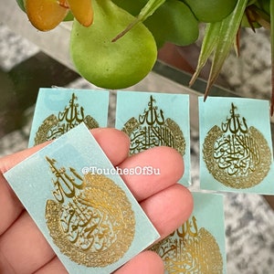 May include: Four blue square stickers with gold Arabic calligraphy. The calligraphy is in a circular pattern with the word "Allah" in the center. The stickers are on a table with a green plant in the background.