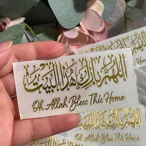 May include: Gold foil stickers with Arabic calligraphy and the English phrase "Oh Allah Bless This Home." The stickers are rectangular and feature ornate script. The background includes greenery and pink flowers.