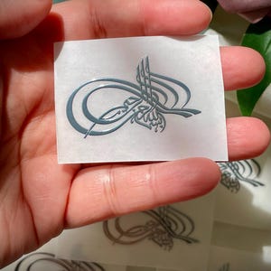 May include: A gray, stylized emblem on a clear sticker. The emblem features ornate, swirling lines and a vertical element with a floral design. Several stickers are visible.