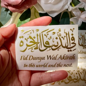 May include: A close-up of a sticker with gold Arabic calligraphy and the English text "Fid Dunya Wal Akirah, In this world and the next." The sticker is held in a hand, with a floral background of white and pink roses.