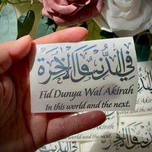 May include: A rectangular sticker with Arabic calligraphy and the English translation "Fid Dunya Wal Akirah, In this world and the next." The text is in a metallic silver color. The background features roses.