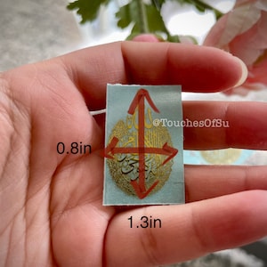 May include: A light blue sticker with gold Arabic calligraphy and a red arrow design. The sticker is 1.3 inches wide and 0.8 inches tall. @TouchesOfSu