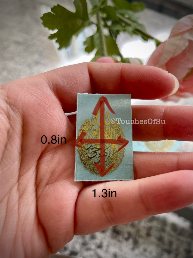 May include: A light blue sticker with gold Arabic calligraphy and a red arrow design. The sticker is 1.3 inches wide and 0.8 inches tall. @TouchesOfSu
