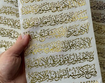May include: Gold foil calligraphy stickers with Arabic script on a white background. The stickers have a glossy finish and are stacked, with some held by a hand. The script is in a flowing, ornate style.