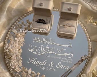 May include: A circular mirrored tray decorated with white floral accents and pearls, featuring two cream-colored ring boxes holding a black band and a gold ring. The mirror displays Arabic calligraphy, the text "And We created you in pairs. Quran 78:8", "Haydr & Sara", and "May 2, 2026", alongside a decorative pen.
