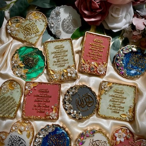 May include: A collection of decorative coasters in various shapes, including hearts, squares, and circles. The coasters feature ornate designs with gold accents, floral embellishments, and Arabic calligraphy. Colors include white, pink, green, blue, and gray.