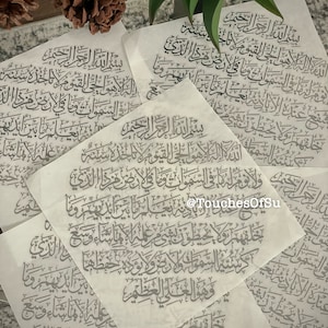 May include: Several sheets of translucent paper with Arabic calligraphy in gray ink. The text is arranged in a flowing script, with multiple sheets overlapping. The background includes pine cones and green leaves.