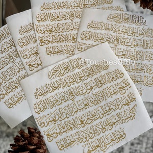 May include: Multiple white sheets of paper with gold Arabic calligraphy printed on them. The sheets are stacked on top of each other and the calligraphy is visible on all of the sheets.