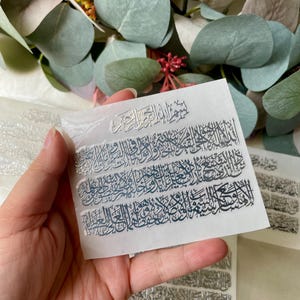 May include: Silver foil stickers featuring Arabic calligraphy. The stickers are on a white backing and are arranged in rows. The calligraphy is detailed and appears to be religious text. The background includes green and pink foliage.