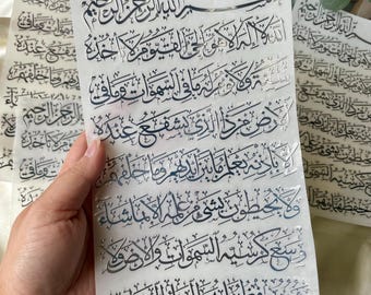 May include: Several white sheets with silver Arabic calligraphy are held in a hand. The sheets are stacked, and the text is visible on each one. The calligraphy is detailed and ornate, with a traditional script.