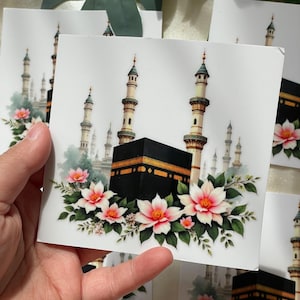 May include: White cards featuring a detailed illustration of the Kaaba, surrounded by minarets and pink and white flowers. The cards have a square shape and a glossy finish, suitable for various occasions.