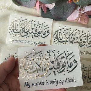 May include: Rectangular stickers with Arabic calligraphy in silver and gold tones. The text "My success is only by Allah" is printed below the calligraphy. The stickers are displayed against a backdrop of green and pink floral elements.