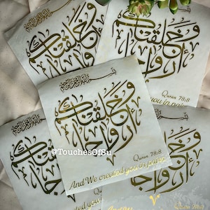 May include: Four white stickers with gold Arabic calligraphy and the text "Quran 78:8 And We created you in pairs." The stickers are on a beige surface.