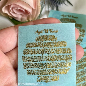 May include: A light blue card with gold lettering that reads "Ayat Ul Kursi" and contains Arabic calligraphy. The card is held in a hand. The text "@TouchesOfSu" is at the bottom of the card.