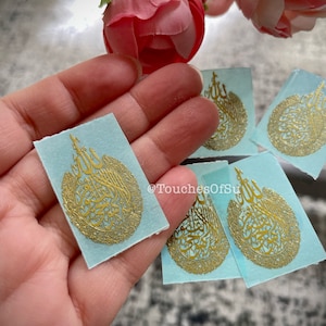 May include: Five small, light blue square stickers with gold Arabic calligraphy in a circular design. The stickers are being held in a hand.
