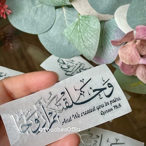 May include: A silver sticker with Arabic calligraphy and the text "And We created you in pairs. Quran 78:8" printed in black. The sticker is on a white background.