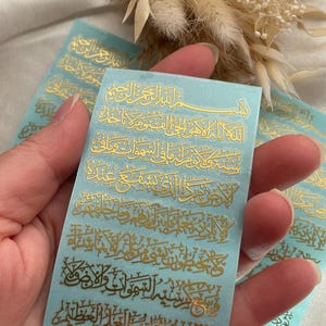 May include: A close-up of several light blue stickers with gold Arabic calligraphy. The stickers are rectangular and feature multiple lines of text. The calligraphy is ornate and detailed, with a shiny, metallic appearance. The stickers are held in a hand.