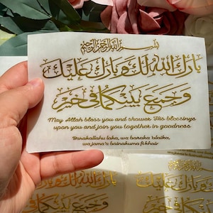 May include: A white card with gold Arabic calligraphy and English text. The text reads "May Allah bless you and shower His blessings upon you and join you together in goodness." The card is held in front of a floral background.