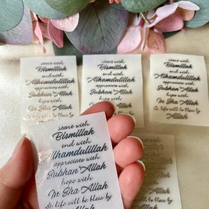 May include: Several small, translucent labels with elegant calligraphy in a script font. The labels feature phrases in a foreign language, with the words "Bismillah", "Alhamdulillah", "Subhan Allah", and "In Sha Allah" visible. The labels are held in a hand.