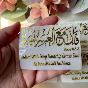 May include: A gold foil sticker with Arabic calligraphy and English text. The sticker reads "Indeed With Every Hardship Comes Ease" and "Fa Inna Ma'al Usri Yusra" from Quran 94:5-6. The sticker is held against a backdrop of pink and green flowers.