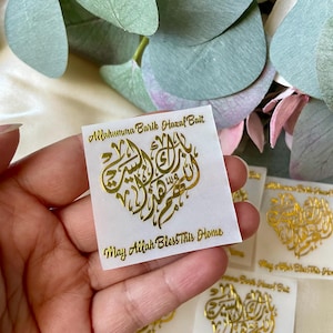 May include: A hand holding several white rectangular stickers with gold calligraphy in a heart shape. The stickers have the text "Allahumma Barik Hazal Bait" and "May Allah Bless This Home". Green and pink foliage is in the background.