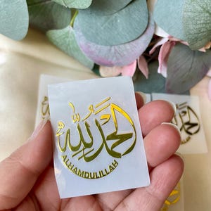 May include: A gold foil sticker with Arabic calligraphy that reads "Alhamdulillah" on a white background. The sticker is held in a hand, with other similar stickers visible in the background. The image is well-lit, with a focus on the sticker's details.