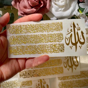 May include: Gold foil stickers featuring Arabic calligraphy. The stickers have a rectangular shape with intricate script and a stylized word. The background includes pink and white flowers.