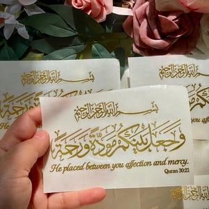 May include: Several white cards with gold calligraphy and the English text "He placed between you affection and mercy. Quran 30:21". The cards are surrounded by pink and white roses and green leaves.