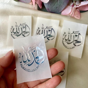 May include: Silver-colored metal stickers with Arabic calligraphy that reads "Alhamdulillah" on small white cards. The stickers are held in a hand, with several others visible in the background. The stickers have a reflective, metallic finish.