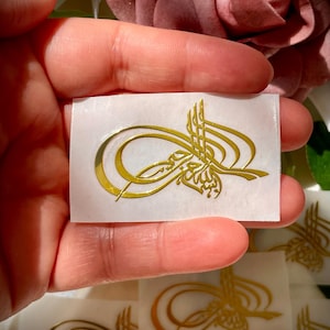 May include: A gold-colored, stylized calligraphy design on a rectangular white sticker. The sticker is held in a hand, with several similar stickers visible in the background. The design features elegant curves and intricate details.