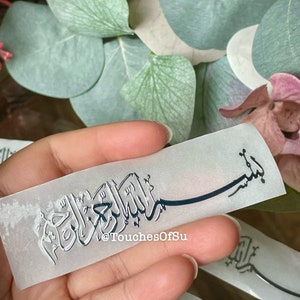 May include: A rectangular, translucent sticker with Arabic calligraphy in a silver metallic finish. The sticker features the text "Bismillah" and the Instagram handle "@TouchesOfSu". The background includes green leaves and a pink flower.