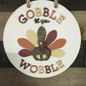 May include: A round wooden sign with the words "Gobble til you Wobble" in brown, orange, and yellow letters. A cutout turkey with brown, orange, and yellow feathers is in the center of the sign.