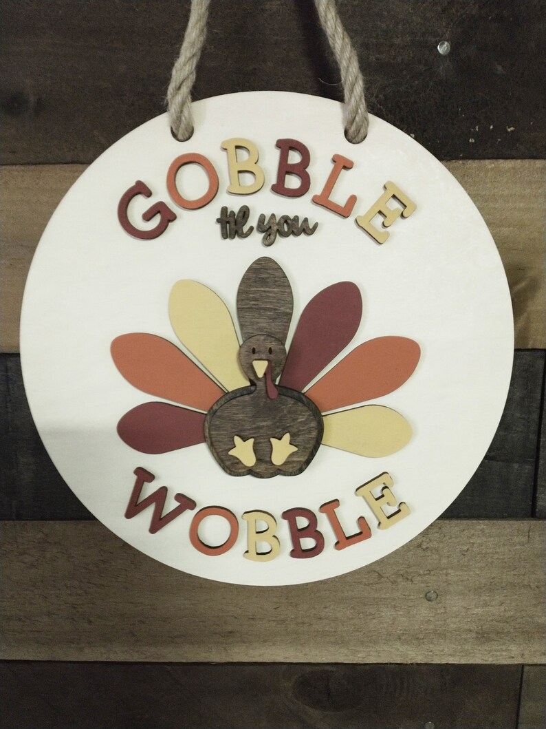 May include: A round wooden sign with the words "Gobble til you Wobble" in brown, orange, and yellow letters. A cutout turkey with brown, orange, and yellow feathers is in the center of the sign.