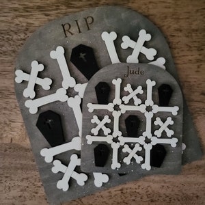 May include: A grey wooden tic-tac-toe game with white bone-shaped pieces and black coffin-shaped pieces. The game board is labelled "RIP" and "Jude".